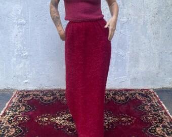 Könnte beinhalten: Ein zweiteiliges burgunderfarbenes Strick-Set, bestehend aus einem Camisole-Top und einem bodenlangen Rock. Das Outfit wird mit weißen Cat-Eye-Sonnenbrillen und Schuhen mit Schlangenmuster getragen. Das Ensemble wird auf einem gemusterten Teppich präsentiert.