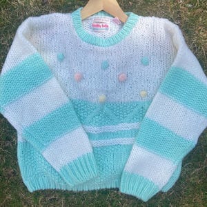 Puede incluir: Un suéter de punto blanco y azul claro con mangas a rayas y cuello redondo. El suéter tiene un ribete verde menta y está decorado con pequeños pompones de colores. La etiqueta dice "knitty bits".