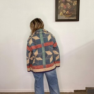May include: A colorful patterned jacket with a geometric design in shades of blue, red, and beige. The jacket has a collar and is worn with blue jeans. The jacket's design features star-like shapes and vertical stripes.