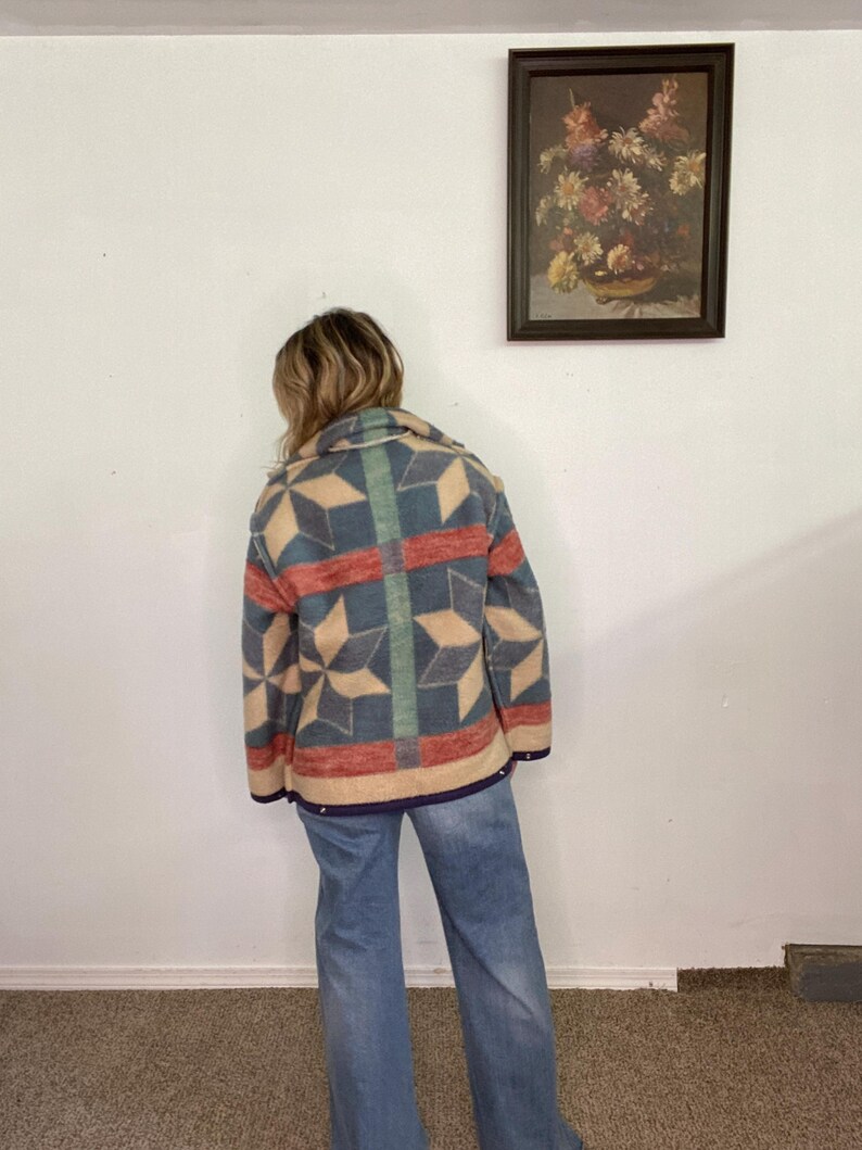 May include: A colorful patterned jacket with a geometric design in shades of blue, red, and beige. The jacket has a collar and is worn with blue jeans. The jacket's design features star-like shapes and vertical stripes.