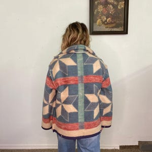 May include: A vintage-style jacket with a geometric star pattern in shades of blue, red, and beige. The jacket has a collared neckline and a button-up front. The jacket is worn with blue jeans.