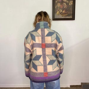 May include: A patterned jacket in shades of blue, purple, and peach. The jacket features a geometric design with star-like shapes and a high collar. The bottom hem is a deep purple color. The person is wearing light blue jeans.