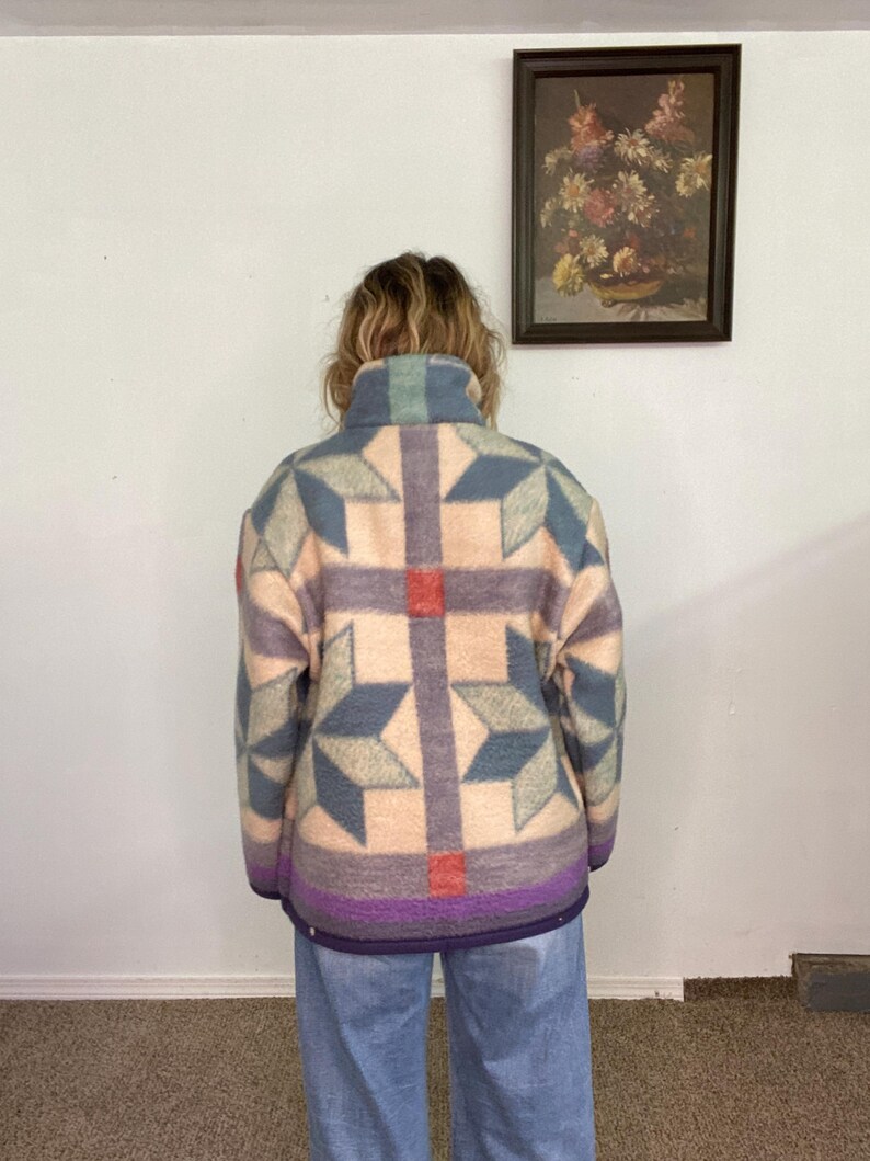May include: A patterned jacket in shades of blue, purple, and peach. The jacket features a geometric design with star-like shapes and a high collar. The bottom hem is a deep purple color. The person is wearing light blue jeans.