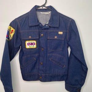 Puede incluir: Chaqueta vaquera azul vintage con cuello, cierre de botones y múltiples bolsillos. La chaqueta presenta parches con texto y gráficos, incluido "That's a BIG 10-4". La chaqueta está colgada de una percha negra.