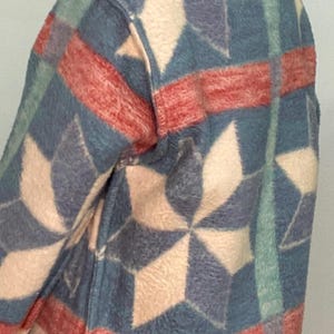 May include: A patterned jacket featuring a geometric design in shades of blue, red, and cream. The jacket has a soft, textured appearance and a dark blue trim along the bottom edge. The design includes star-like shapes.