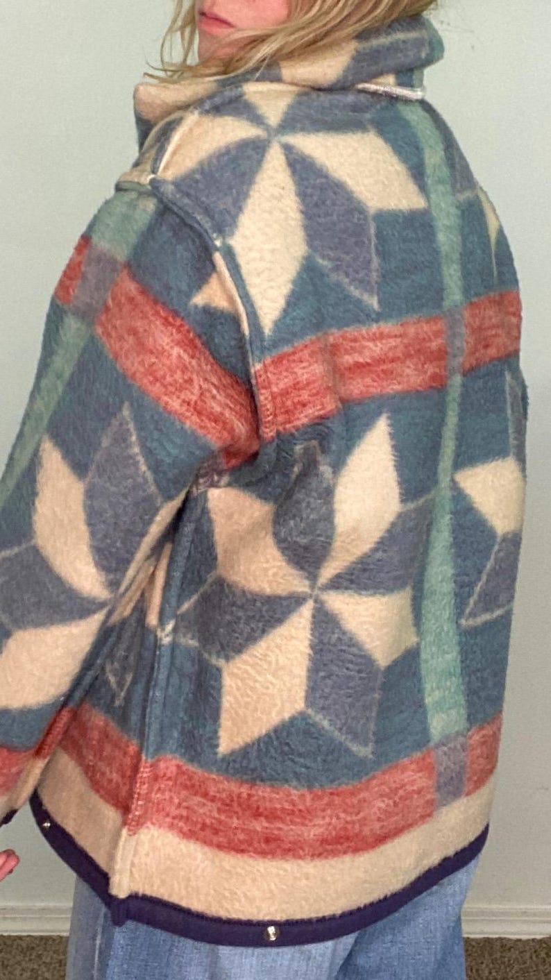 May include: A patterned jacket featuring a geometric design in shades of blue, red, and cream. The jacket has a soft, textured appearance and a dark blue trim along the bottom edge. The design includes star-like shapes.