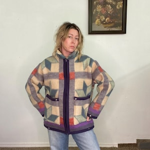 May include: A vintage zip-up jacket with a geometric pattern in beige, gray, teal, and red. The jacket has a purple zipper, trim, and pockets. The model is wearing blue jeans.