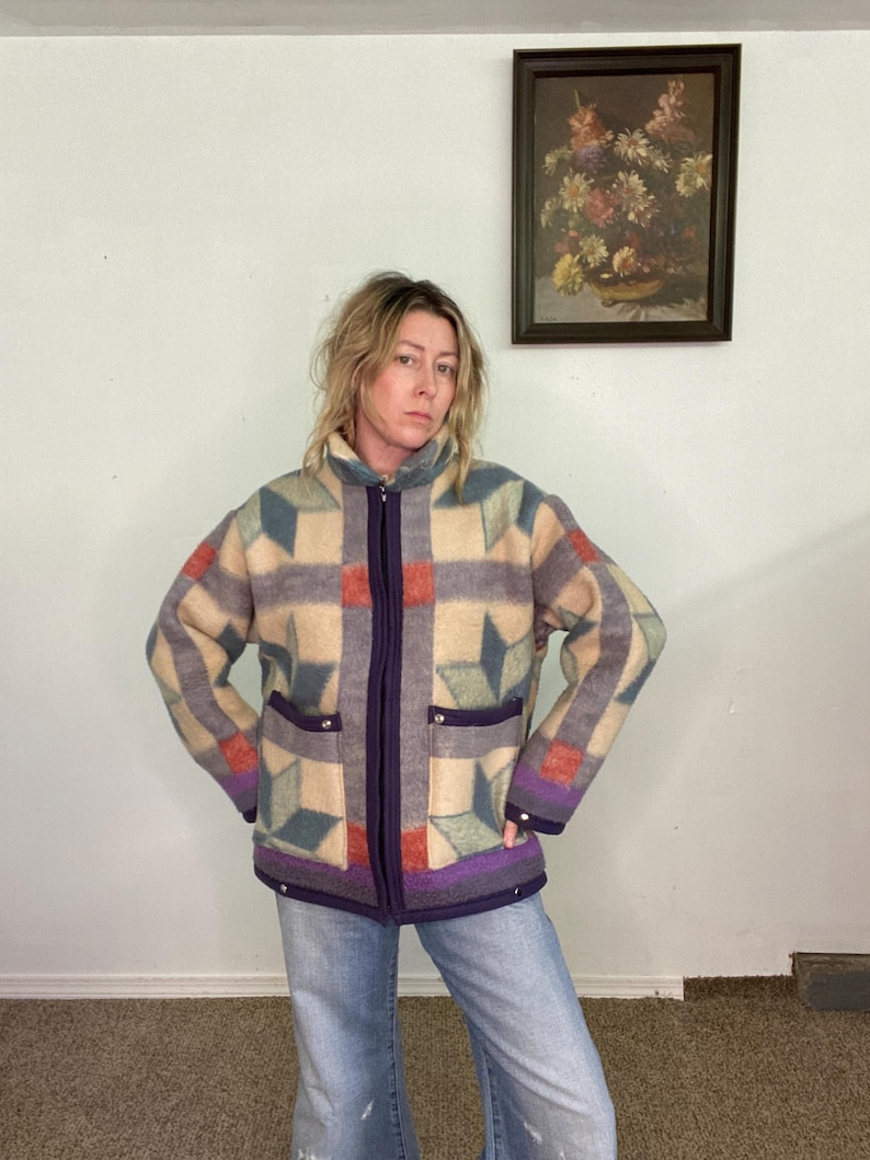 May include: A vintage zip-up jacket with a geometric pattern in beige, gray, teal, and red. The jacket has a purple zipper, trim, and pockets. The model is wearing blue jeans.