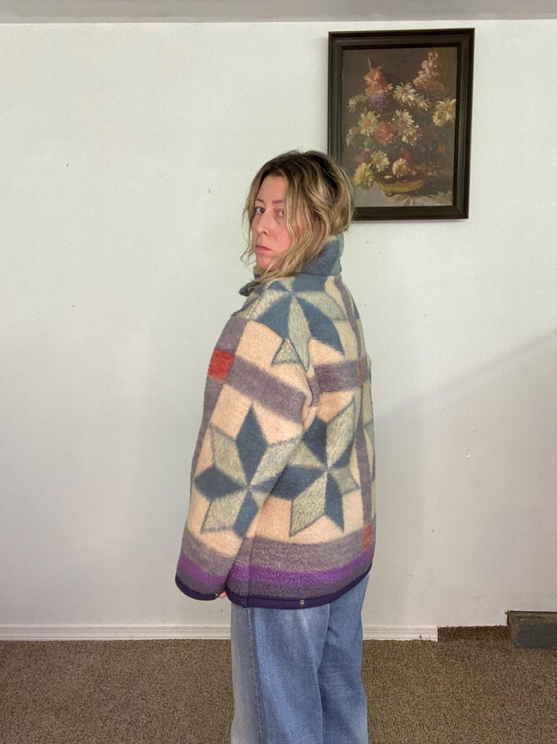 May include: A quilted jacket with a geometric star pattern in shades of blue, purple, and beige. The jacket has a high collar and a purple hem. The jacket is worn with blue jeans.