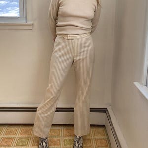 May include: A cream-colored long-sleeved shirt and matching pinstripe pants with a belt. The pants have vertical black stripes. The outfit is paired with patterned ankle boots. The outfit is worn in a room with a patterned floor.
