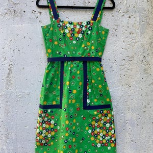 Może przedstawiać: Zielona, ​​zabytkowa sukienka letnia we wzór kwiatowy, z kwadratowym dekoltem, granatowymi wykończeniami i ramiączkami. Sukienka ma dwie przednie kieszenie i granatowy pasek. Sukienka wisi na czarnym wieszaku.