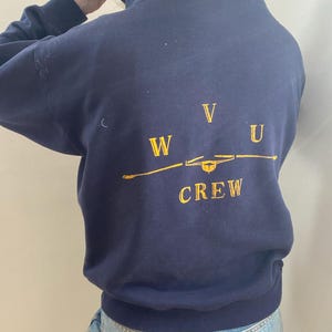 Puede incluir: Sudadera con capucha azul marino con texto dorado bordado en la espalda. El texto dice "WVU CREW" con un gráfico estilizado. La sudadera tiene una capucha con cordón y mangas largas. La persona lleva vaqueros azules.