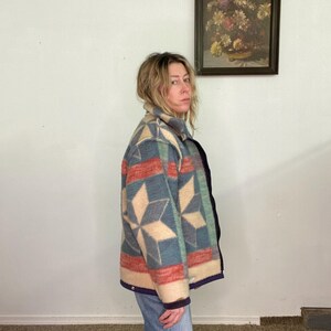 May include: A patterned jacket in shades of red, blue, and beige. The jacket features a geometric design with a collar and a dark blue trim. The person is wearing blue jeans. A framed floral painting hangs on the white wall.