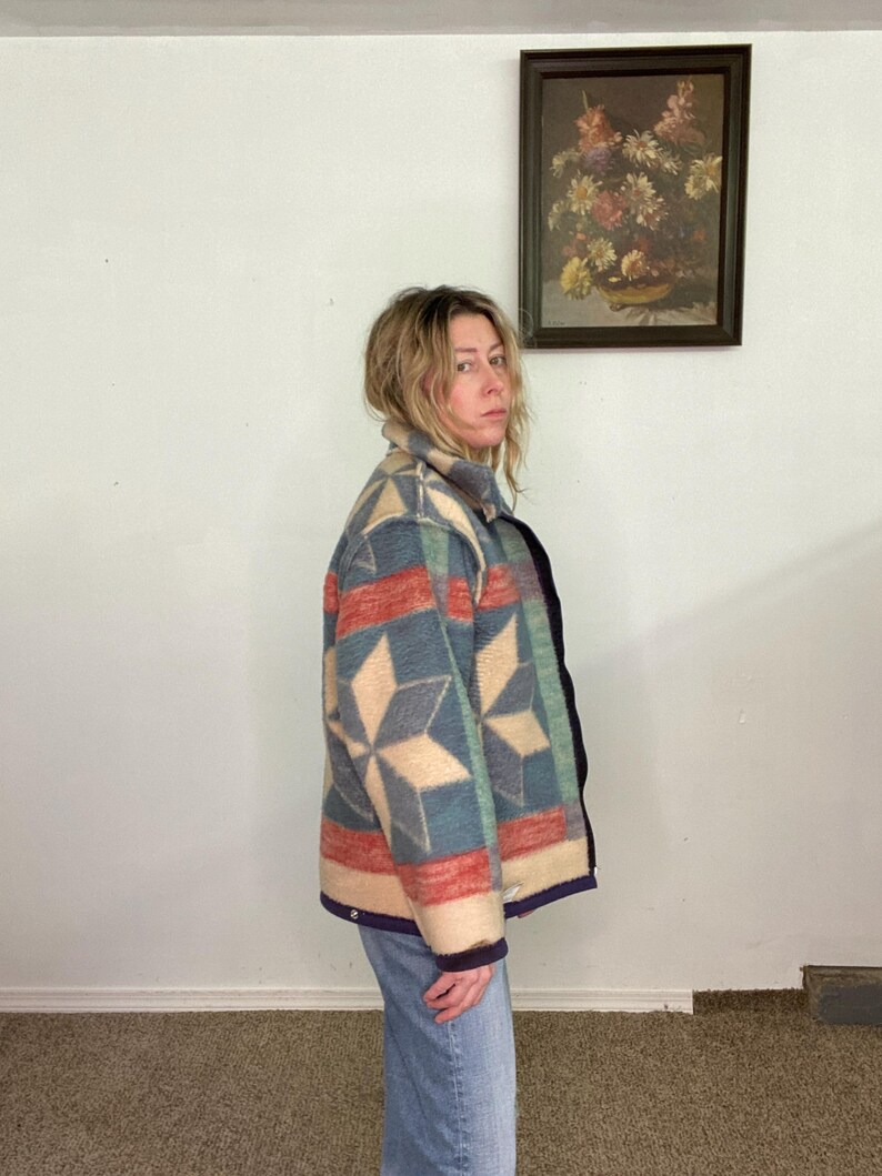 May include: A patterned jacket in shades of red, blue, and beige. The jacket features a geometric design with a collar and a dark blue trim. The person is wearing blue jeans. A framed floral painting hangs on the white wall.