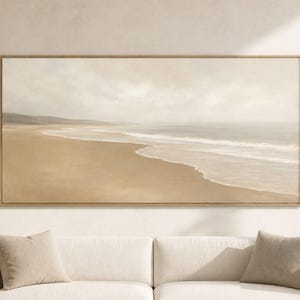 Puede incluir: Un cuadro de paisaje rectangular que muestra una serena escena costera con una playa de arena, suaves olas del mar y un cielo nublado en tonos beige y crema, expuesto en un marco de madera fino sobre un sofá de color crema.