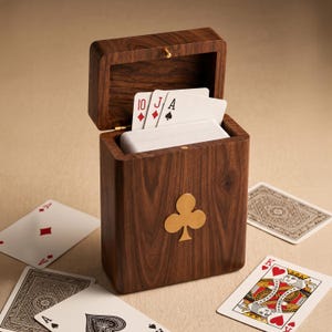 May include: A dark brown wooden playing card box with a hinged lid, featuring a gold-colored metal clover emblem on the front. The box is filled with a deck of playing cards, with the ten of diamonds, jack of diamonds, and ace of spades visible.