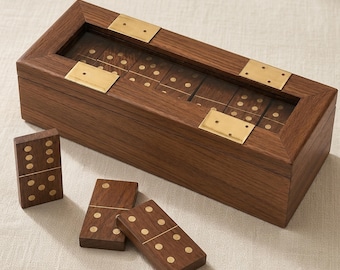 Könnte beinhalten: Eine rechteckige Dominobox aus dunkelbraunem Holz mit Glasdeckel und Messingbeschlägen an den Ecken, die ein Set aus Holzdominos enthält. Drei weitere Holzdominos mit Messingpunkten liegen vor der Box auf einer hellen Oberfläche.