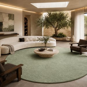 May include: A large, round, sage green area rug with a subtle, textured surface pattern. The rug is positioned in the centre of a room, serving as a foundation for a curved white sofa, a round stone coffee table, and two dark wood armchairs.