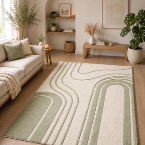 May include: A cream-coloured rectangular rug featuring a design of sage green curved lines and arches. The rug is in a living room setting, alongside a sofa, plants, and wooden furniture. The rug's dimensions are approximately 200cm x 300cm.