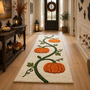 Op de afbeelding: Een rechthoekige, gebroken witte loper met een getuft ontwerp van een kronkelende groene wijnstok met drie oranje pompoenen van verschillende groottes. Het kleed heeft een getextureerd oppervlak en ligt op een lichte houten vloer.