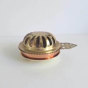 May include: A round brass incense burner featuring a domed lid with vertical cutouts, a copper-colored middle band, and a small decorative handle extending from the side. The object has a polished metallic finish and a circular base structure.