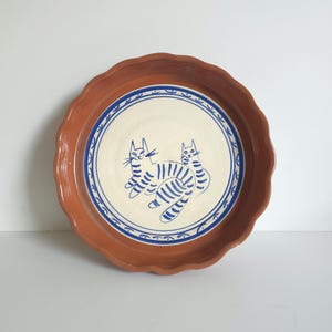 May include: A round terracotta baking dish with a scalloped edge. The interior features a cream-colored circular base decorated with two blue hand-painted cats and a blue border pattern consisting of small arches and dots.