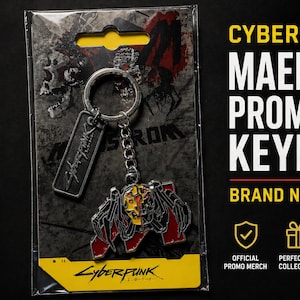 May include: Cyberpunk 2077 Maelstrom promotional keyring. The keyring features a metal design with red, black, and yellow accents. The keyring is attached to a silver chain and a rectangular tag. The packaging includes the text "Cyberpunk 2077".