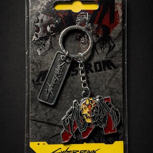 May include: Cyberpunk 2077 keychain featuring a stylized spider-like creature with red and yellow accents. The keychain includes a metal tag with the game's logo. The packaging is a clear plastic with a yellow label and the game's title.
