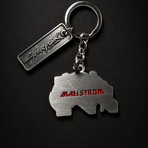 May include: A silver-colored keychain with two metal tags. One tag reads "Cyberpunk" in stylized lettering. The other tag is shaped like a map with the word "Maelstrom" in red lettering. The keychain is on a black background.