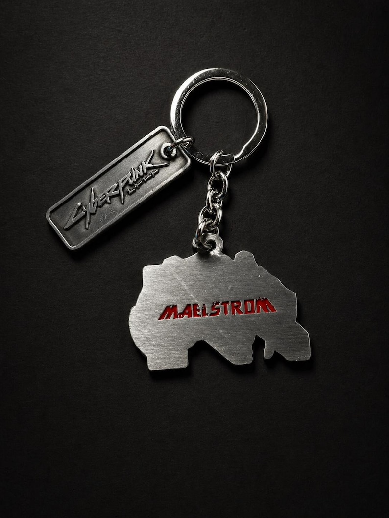 May include: A silver-colored keychain with two metal tags. One tag reads "Cyberpunk" in stylized lettering. The other tag is shaped like a map with the word "Maelstrom" in red lettering. The keychain is on a black background.