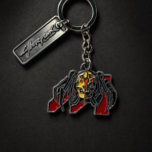 May include: Silver keychain with a rectangular tag featuring the word "Cyberpunk" in a stylized font. The keychain includes a detailed, colorful pendant depicting a spider-like creature with a yellow and red design.