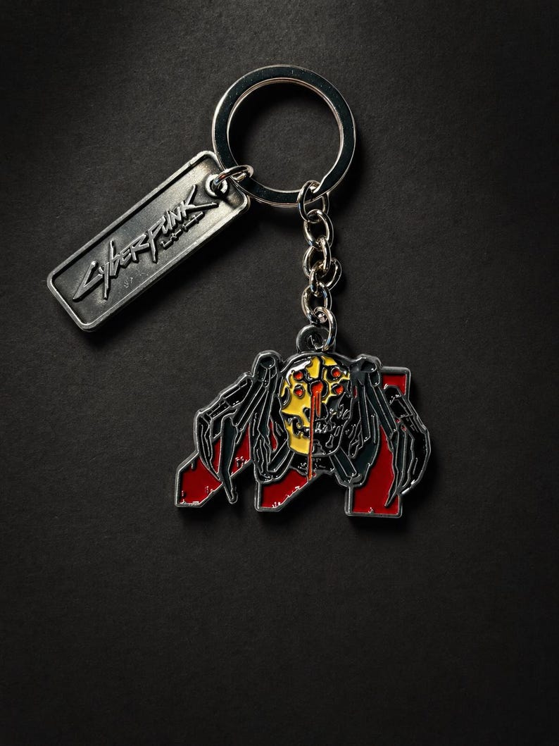 May include: Silver keychain with a rectangular tag featuring the word "Cyberpunk" in a stylized font. The keychain includes a detailed, colorful pendant depicting a spider-like creature with a yellow and red design.