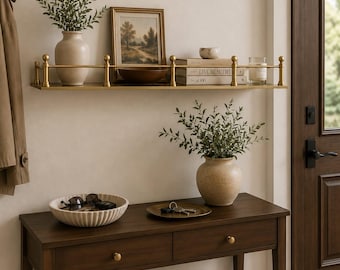 May include: A gold metal wall shelf with a railing, holding a beige vase with greenery, a framed landscape print, a wooden bowl, two books, and a glass candle. Below, a dark wood console table holds a ribbed bowl, a tray with keys, and a vase.