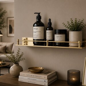 May include: A gold-toned metal wall shelf with a protective rail, holding a large Aesop hand wash bottle, a Meraki dropper bottle, a Meraki body butter jar, and a small green plant in a light-colored ceramic pot against a neutral wall.