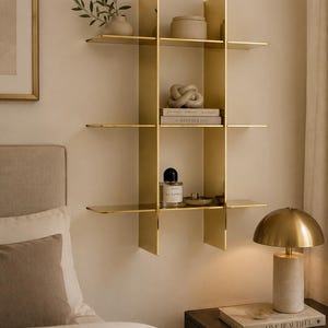 May include: A wall-mounted gold metal shelving unit featuring three horizontal tiers divided by two vertical panels. The shelves hold various decorative items including a small plant, a ceramic container, a knot sculpture, books, and a glass perfume bottle.