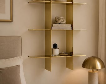 May include: A wall-mounted gold metal shelving unit featuring three horizontal tiers divided by two vertical panels. The shelves hold various decorative items including a small plant, a ceramic container, a knot sculpture, books, and a glass perfume bottle.