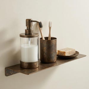 May include: A bronze-toned bathroom shelf holds a soap dispenser, toothbrush holder with two toothbrushes, and a bar of soap on a small dish. The soap dispenser has a clear glass body and a pump. The shelf is mounted on a light-colored wall.