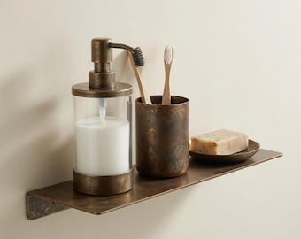 May include: A bronze-effect bathroom shelf with a soap dispenser, toothbrush holder with two toothbrushes, and a bar of soap on a small dish. The soap dispenser has a clear glass body and a pump. The shelf is mounted on a light-coloured wall.