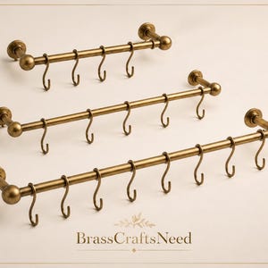 May include: Three antique brass-coloured hook racks. Each rack has a horizontal bar with multiple curved hooks. The racks are mounted on a light-coloured wall. The text "BrassCraftsNeed" is at the bottom.