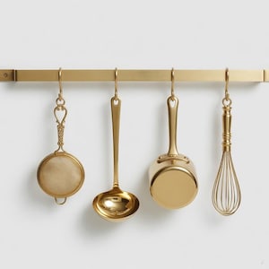 Op de afbeelding: Een gouden keukengerei set hangend aan een bijpassende stang. De set bevat een zeef, een pollepel, een kleine steelpan en een garde. De items hangen tegen een effen witte achtergrond, wat een strakke en elegante uitstraling creëert.