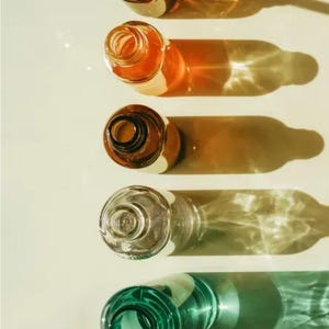 May include: Overhead shot of five glass bottles in a row, each casting a long shadow. The bottles are various colors, including amber, clear, and green. The bottles are empty and have open necks.