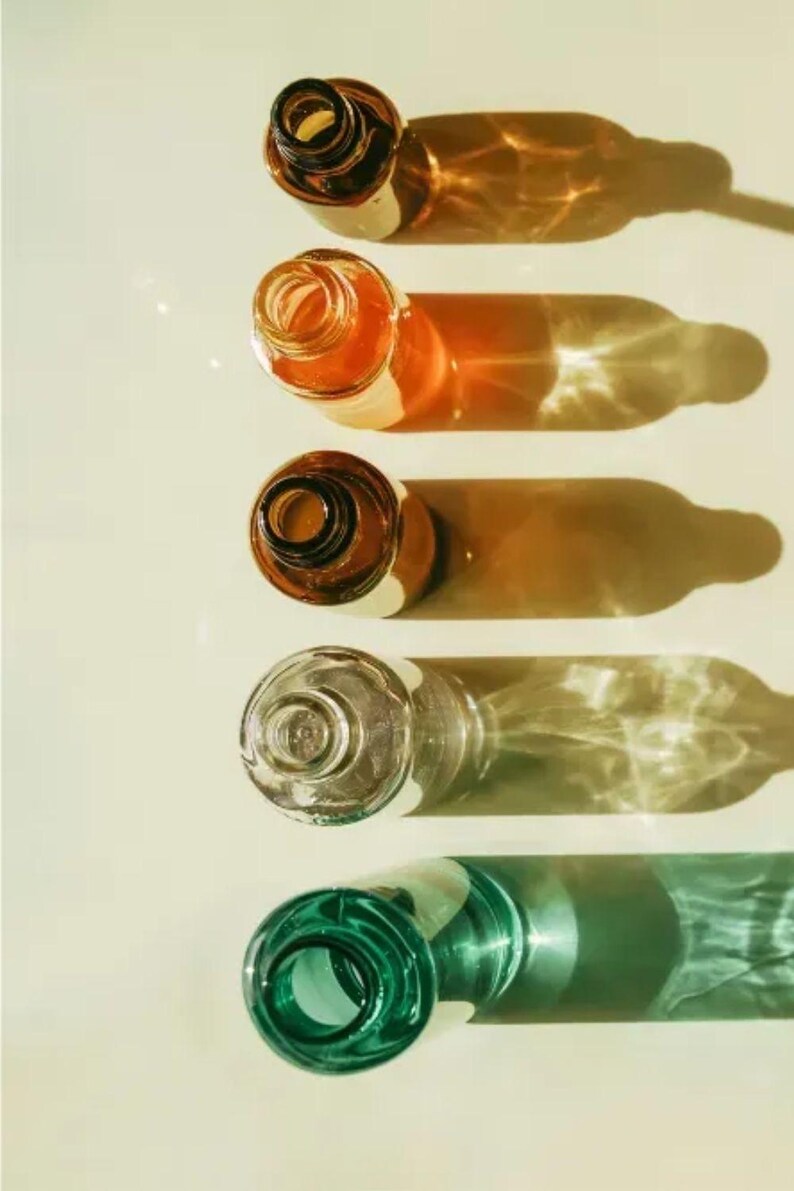 May include: Overhead shot of five glass bottles in a row, each casting a long shadow. The bottles are various colors, including amber, clear, and green. The bottles are empty and have open necks.