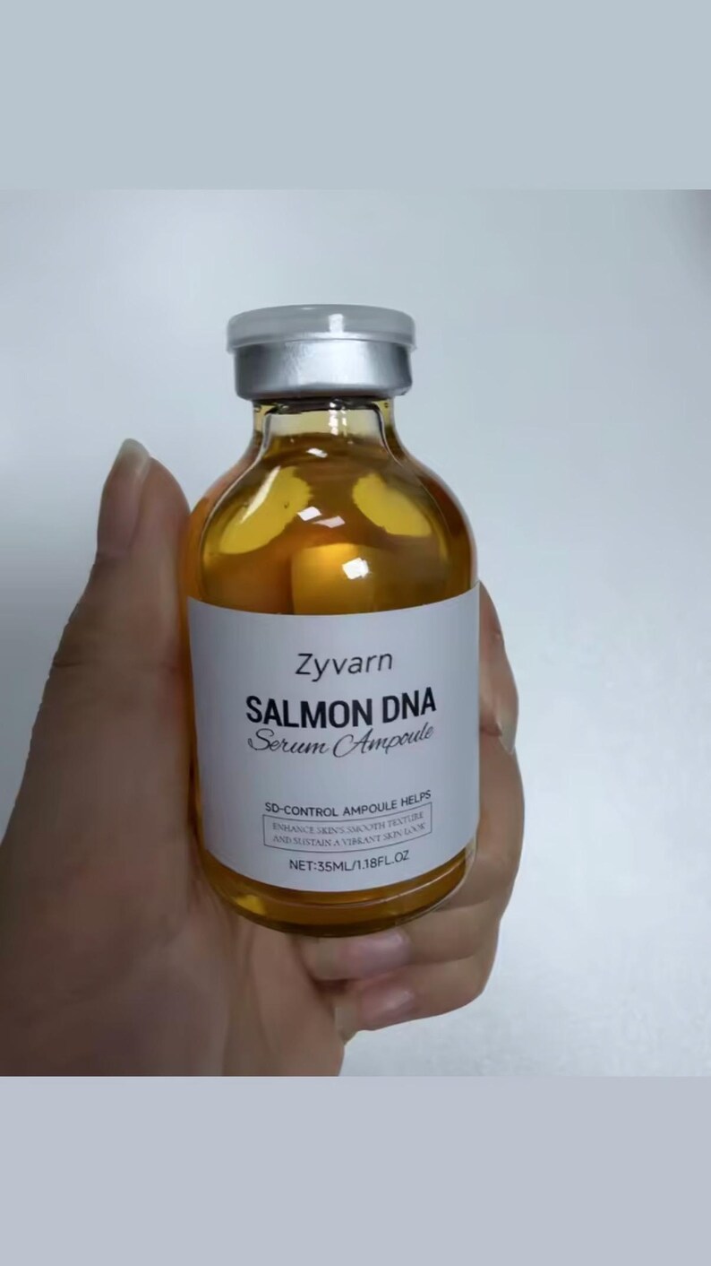 May include: A clear glass bottle of Zyvarn Salmon DNA Serum Ampoule, held in a hand. The bottle has a silver cap and a white label with black text. The label reads "SD-CONTROL AMPOULE HELPS ENHANCE SKIN'S SMOOTH TEXTURE AND SUSTAIN A VIBRANT SKIN GLOW". The bottle contains 35ml/1.18 fl oz.