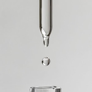 May include: A close-up shot of a clear glass dropper with a single drop of liquid suspended mid-air, above the neck of a clear glass bottle. The bottle and dropper are against a plain white background.