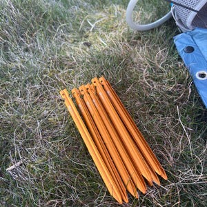 May include: Six orange tent pegs lie on green grass. The pegs are made of metal and have a pointed end for easy ground insertion. They are designed to secure tents and other outdoor equipment.