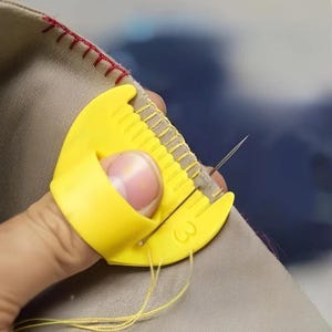 May include: A bright yellow plastic sewing thimble guide worn on the thumb, featuring a series of small grooves along the top edge for spacing stitches. The tool is positioned against beige fabric with red and yellow thread.