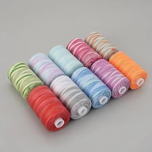 May include: A collection of ten cylindrical spools of sewing thread arranged in two rows. The threads feature variegated color patterns including red, green, light blue, white, grey, pink, purple, and orange, each wound on a white plastic core.