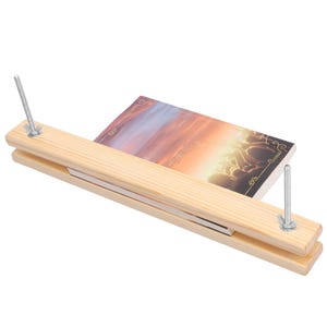 May include: A light-colored wooden bookbinding press featuring two long, rectangular wooden boards held together by two vertical metal threaded rods with adjustable wing nuts, clamping a book with a sunset-themed cover illustration.