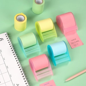 May include: A collection of colorful adhesive note dispensers in pastel pink, yellow, light green, and blue, each holding a roll of paper. One pink note has the handwritten text Creative Post-it Notes with a smiley face. A spiral calendar and pen are nearby.