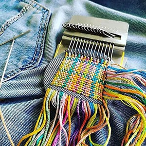 May include: A metal darning loom tool positioned on blue denim fabric, featuring a square woven patch of multicolored thread. The loom has silver hooks holding the warp threads, with loose, vibrant yarn strands extending from the bottom edge.
